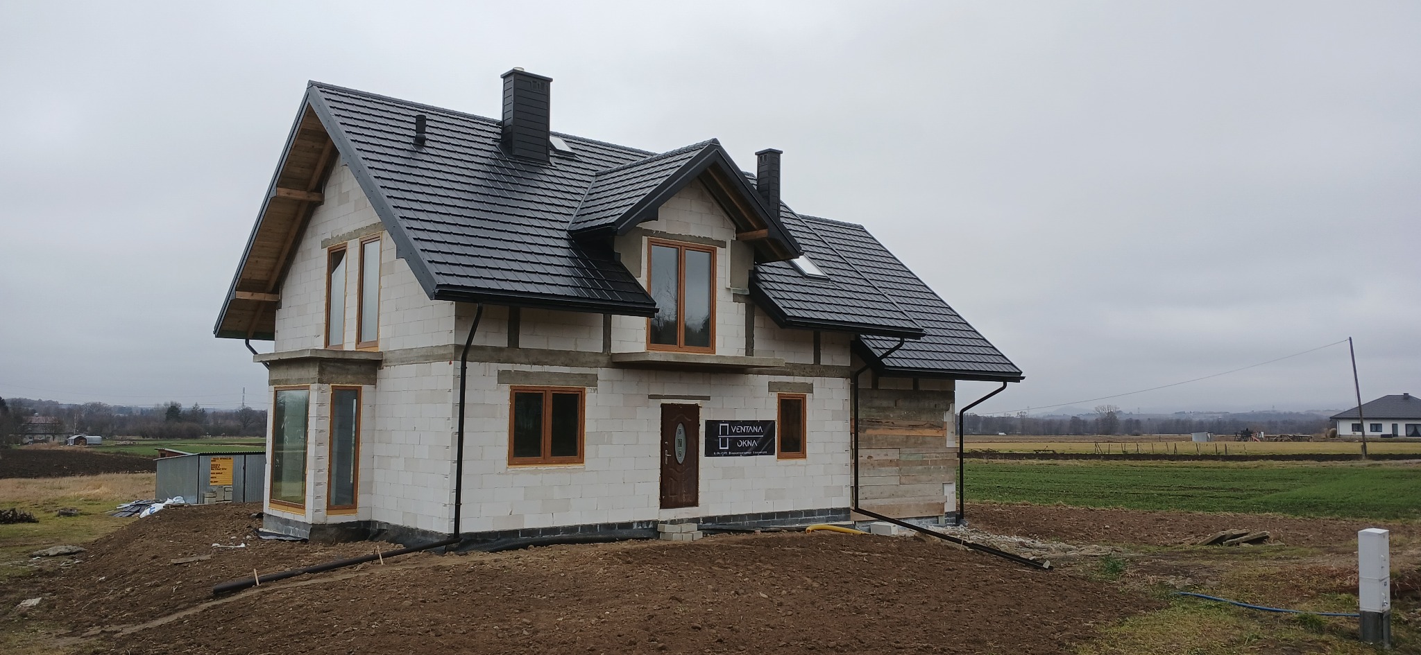 Dom w trakcie budowy z czarną dachówką, białymi ścianami z betonu komórkowego i drewnianymi oknami. Widoczne elementy konstrukcyjne i krajobraz w tle.