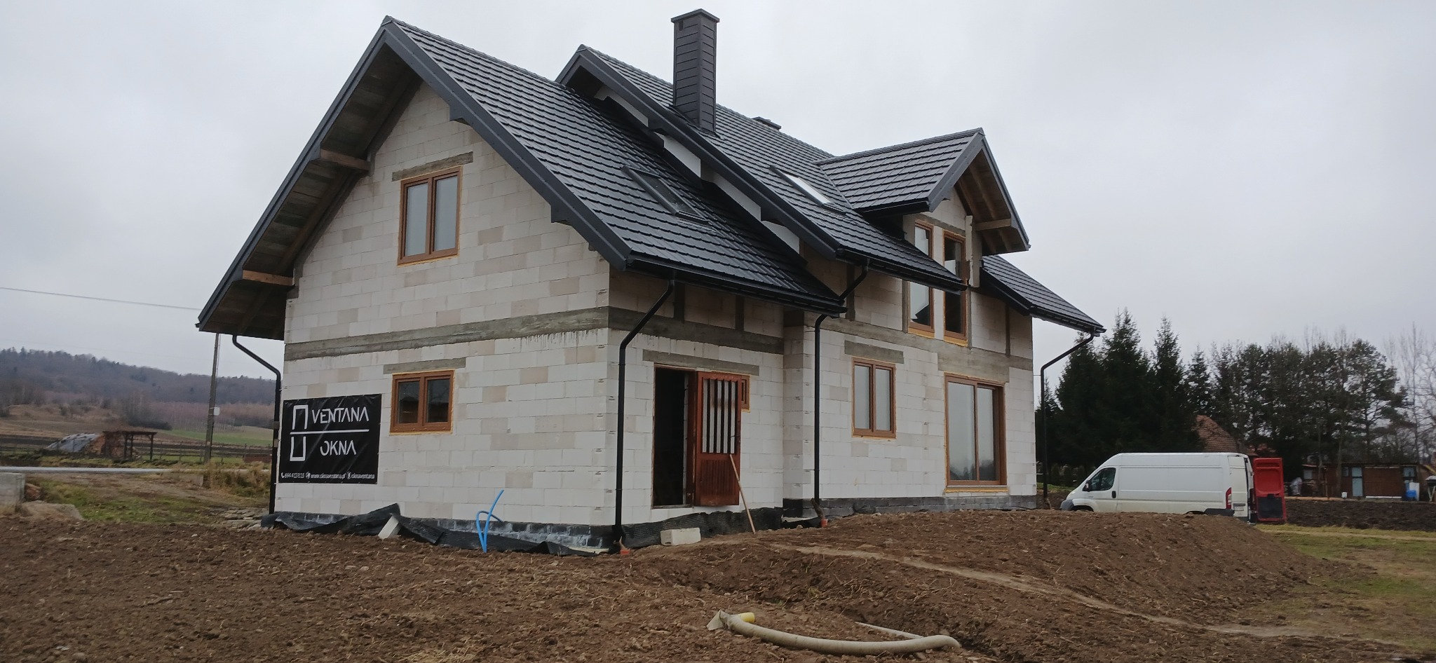 Surowy dom z białego pustaka z czarną dachówką i oknami w drewnianych ramach. Na ścianie baner reklamowy firmy okiennej. W tle zalesione wzgórza i biały van.