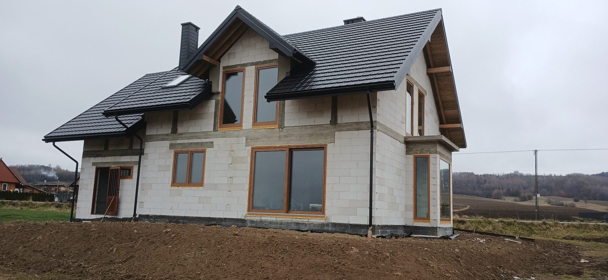 Niedokończony dom z białych bloczków, z czarnym dachem i brązowymi oknami. Widoczne elementy konstrukcyjne i krajobraz w tle. Dom w stanie surowym otwartym.
