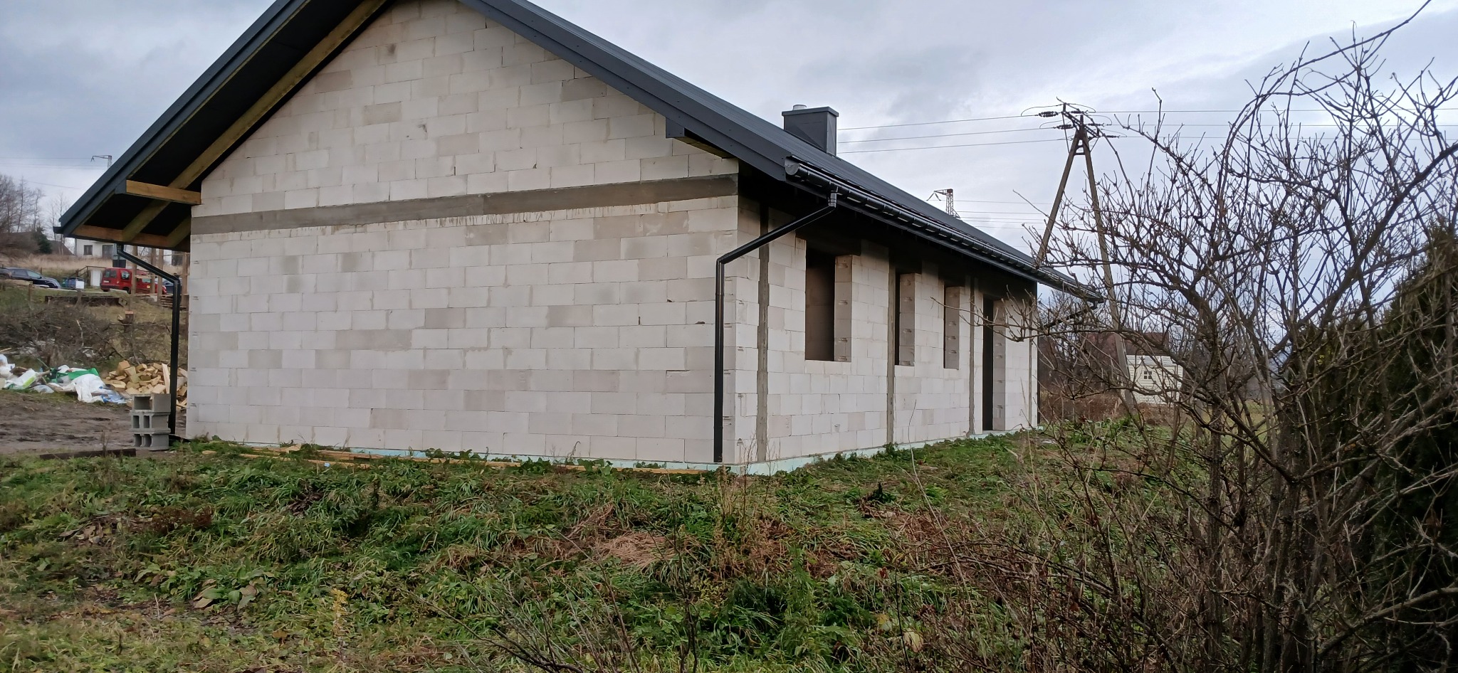 Surowy, parterowy dom z białych bloczków, z czarnym dachem i rynnami, w trakcie budowy. Widoczne otwory okienne i fragmenty instalacji. Tło: pochmurne niebo, krzewy.