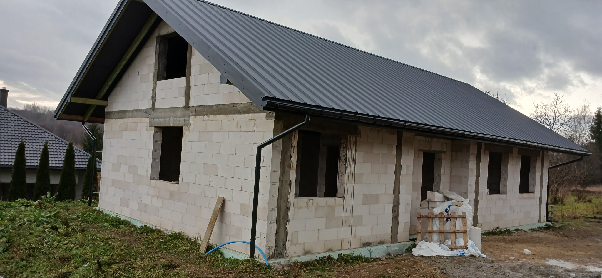 Niedokończony dom z białych bloczków, z szarym dachem z blachy. Widoczne niezabezpieczone otwory okienne i drzwiowe. Na ziemi leżą materiały budowlane.