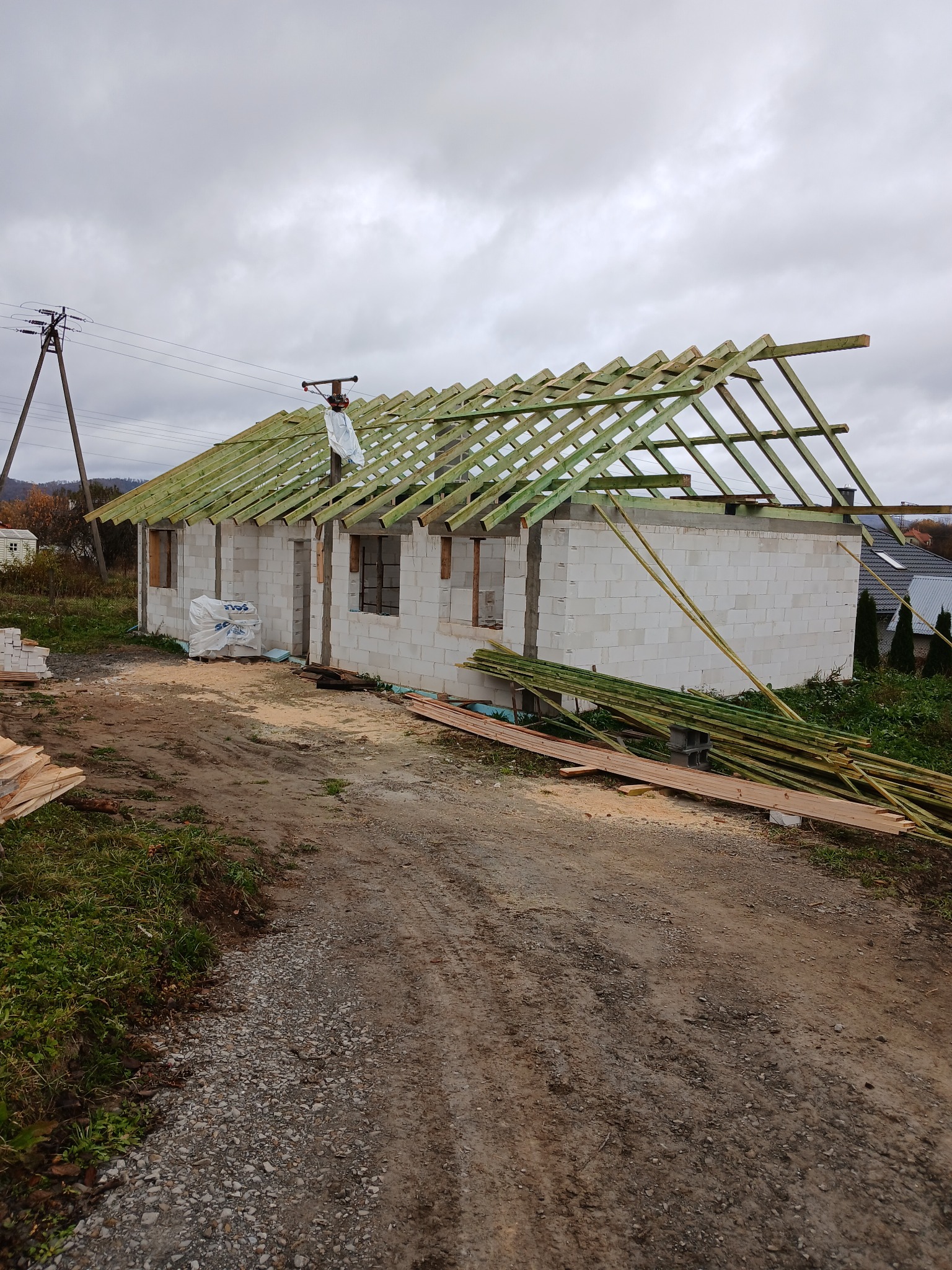 Budowa domu z białych bloczków: ściany wzniesione, konstrukcja dachu z zielonych belek w trakcie montażu, materiały budowlane obok. Pochmurne niebo w tle.