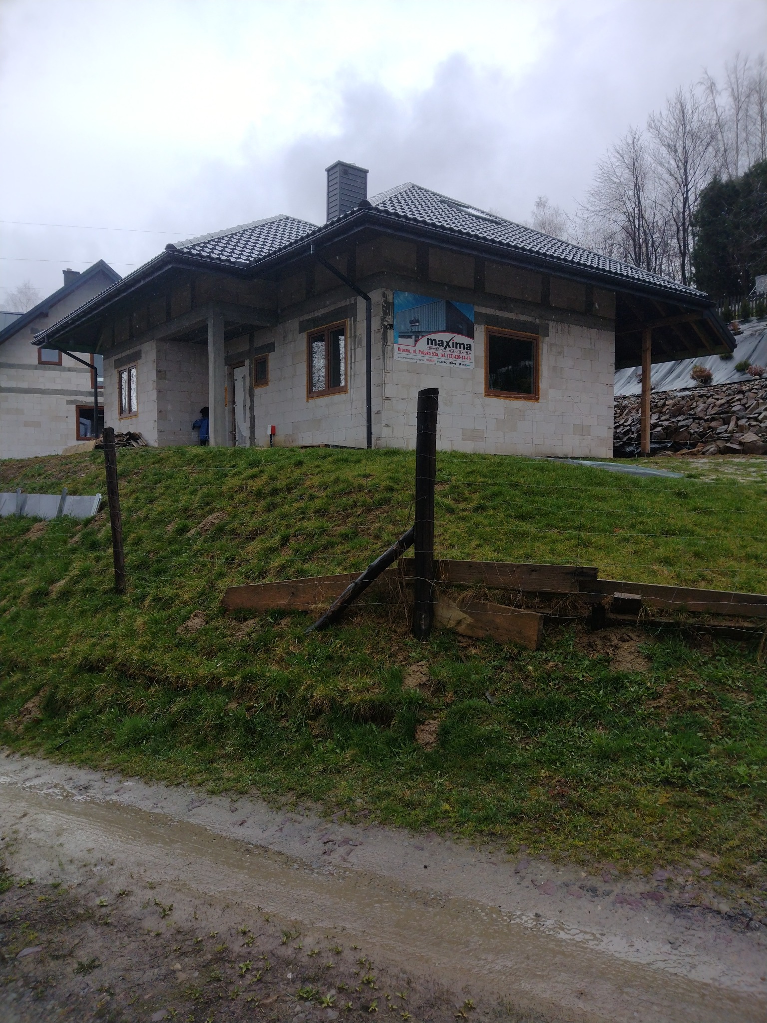 Surowy dom z białego pustaka na wzgórzu, z widocznymi oknami i dachem z dachówki. Na ścianie baner reklamowy. Teren błotnisty, pochmurne niebo.