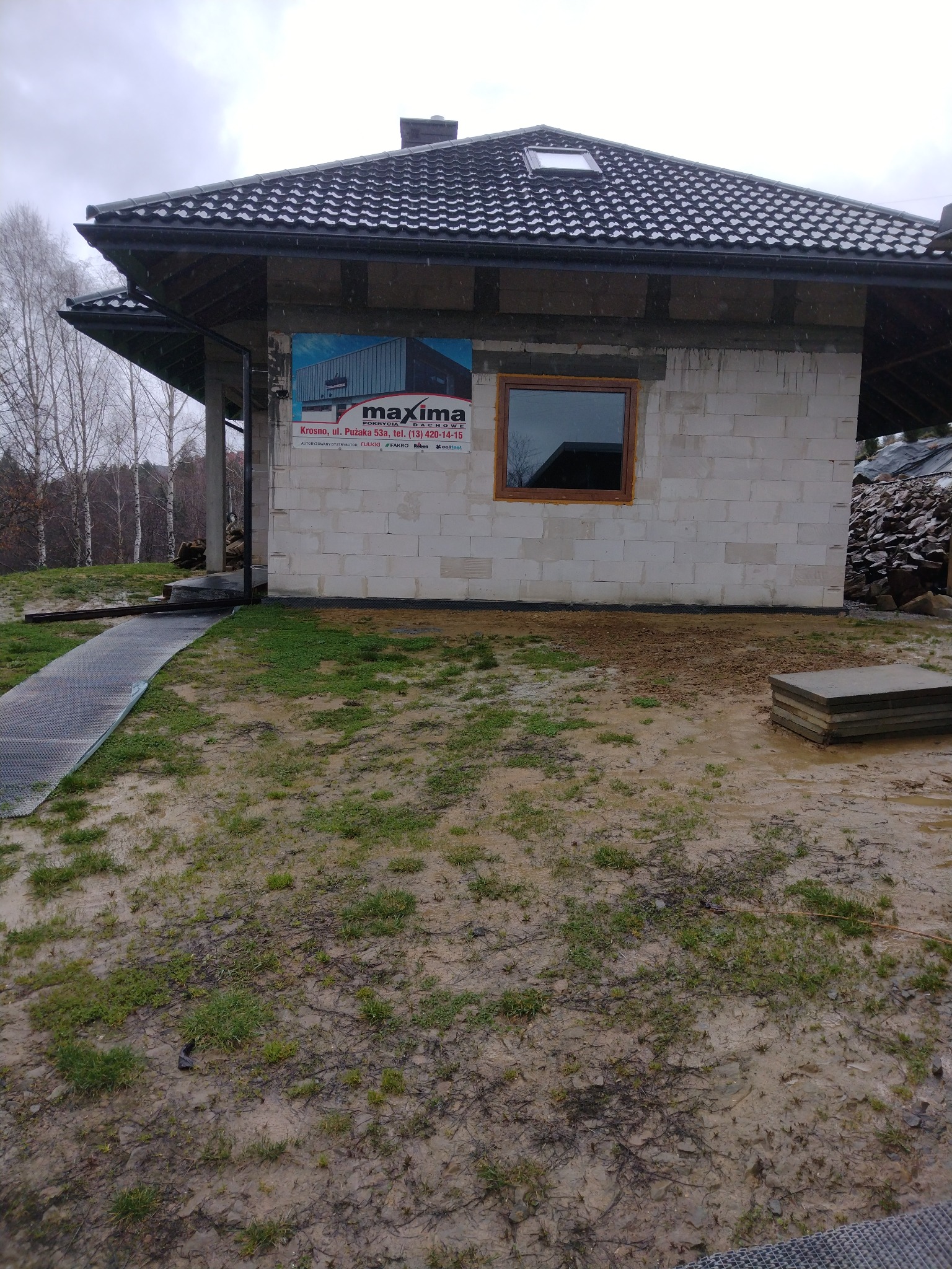 Niedokończony dom z białych bloczków, z oknem w drewnianej ramie i czarnym dachem. Na ścianie reklama firmy Maxima. Teren wokół błotnisty, pochmurna pogoda.