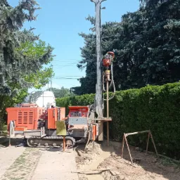 Pomarańczowa wiertnica gąsienicowa w trakcie pracy, widoczne błoto i wykop, tło stanowią drzewa iglaste i żywopłot.