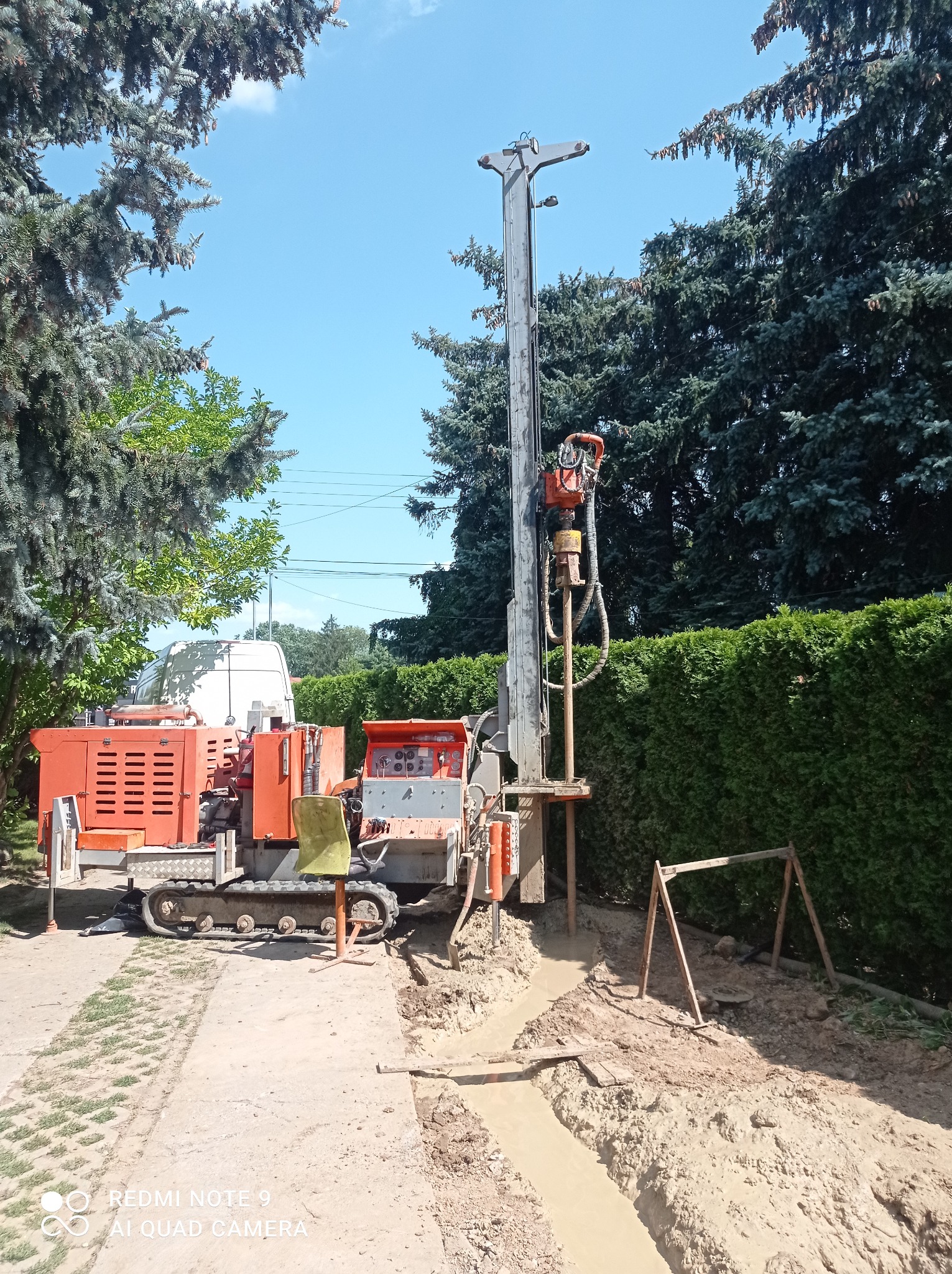 Pomarańczowa wiertnica gąsienicowa w trakcie pracy, widoczne błoto i wykop, tło stanowią drzewa iglaste i żywopłot.