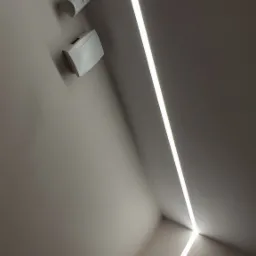 Minimalistyczne oświetlenie LED w rogu pomieszczenia, z czujnikiem ruchu i głośnikiem na ścianie, skierowane ku górze, na tle gładkiej ściany w odcieniach szarości.