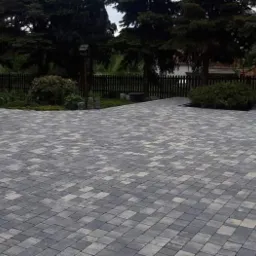 Ułożona szara kostka brukowa w jodełkę na podjeździe, z widocznym ogrodzeniem drewnianym w tle i zielenią ogrodu.