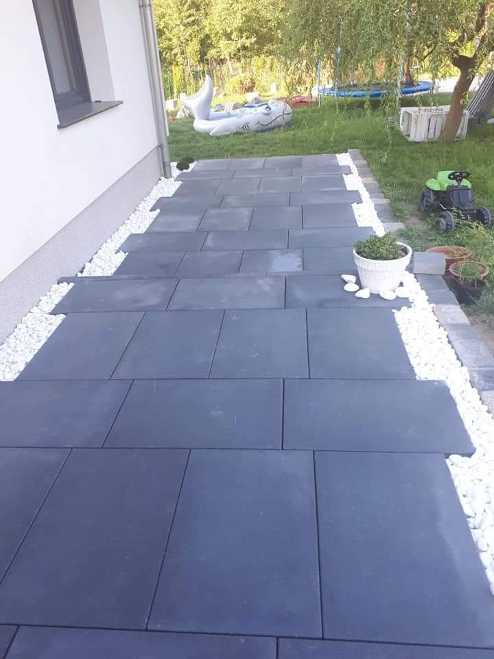 Ścieżka z dużych, grafitowych płyt betonowych, obłożona białymi kamykami, prowadząca przez trawnik z dmuchanym rekinem i zabawkami.