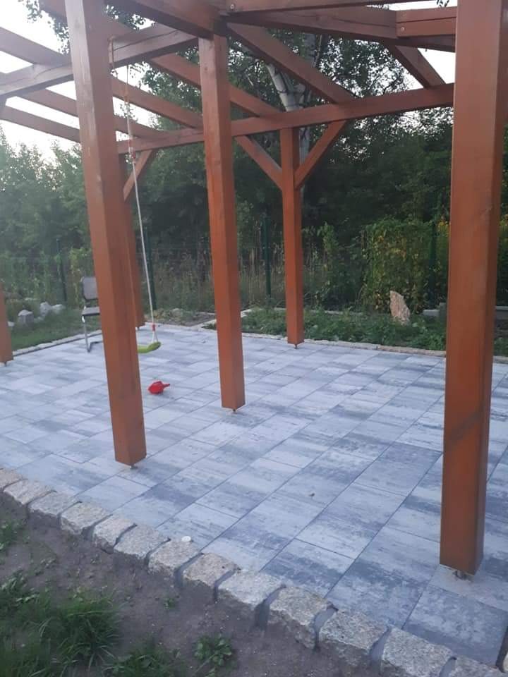 Ułożona kostka brukowa w szaro-niebieskim odcieniu pod drewnianą konstrukcją altany z huśtawką, obrzeże z kostki granitowej, widok na ogród w tle.