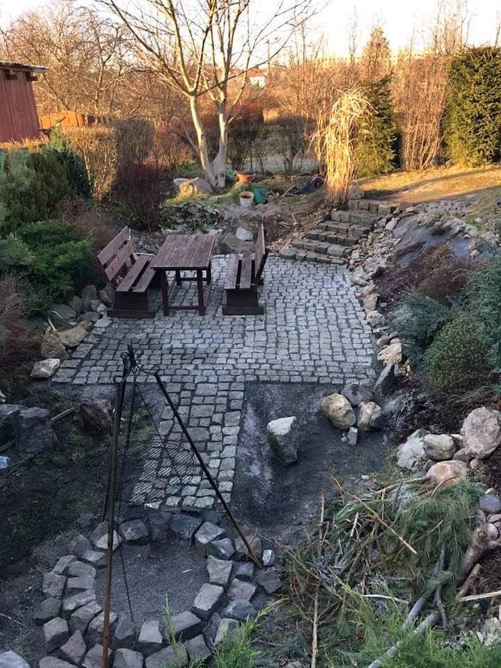 Ułożona kostka na patio z drewnianym zestawem mebli ogrodowych, kamienny krąg paleniska z trójnogiem do grillowania w tle, schody z kamieni prowadzące do ogrodu.