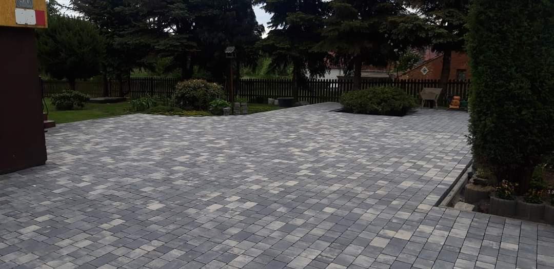 Ułożona szara kostka brukowa w jodełkę na podjeździe, z widocznym ogrodzeniem drewnianym w tle i zielenią ogrodu.