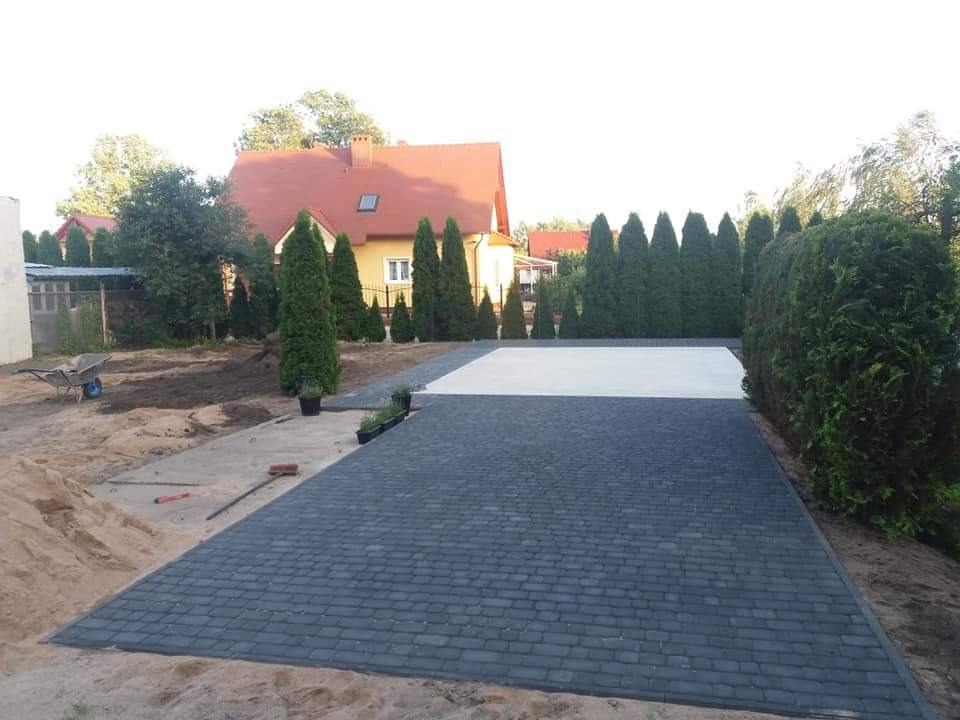 Ułożona ciemnoszara kostka brukowa prowadząca do białej wylewki betonowej, w tle dom z pomarańczowym dachem i rząd tuj, widoczna taczka i przygotowany teren pod dalsze prace.