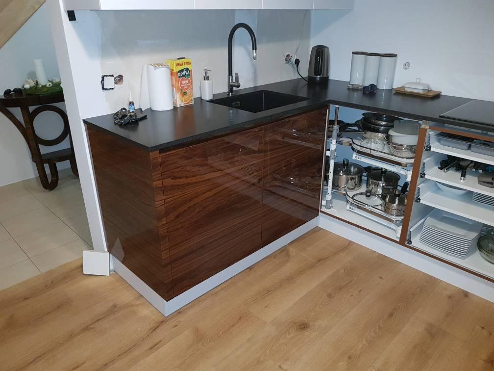 Kuchenny narożnik z ciemnym blatem i frontami szafek z wyraźnym rysunkiem drewna, otwarty regał narożny z garnkami i naczyniami, drewnopodobna podłoga.