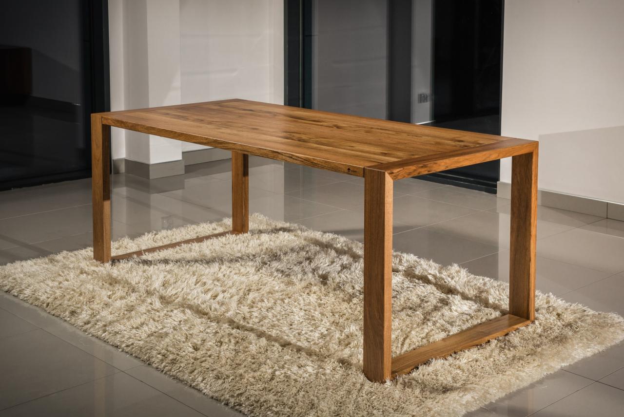Stół z litego drewna o prostej konstrukcji, ustawiony na puszystym, kremowym dywanie w nowoczesnym wnętrzu z szarymi płytkami podłogowymi.