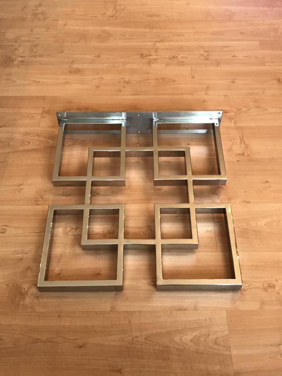 Metalowa konstrukcja z kwadratowych profili, układanka geometryczna, przygotowana do montażu na ścianie, widoczne śruby mocujące, na podłodze imitującej drewno.