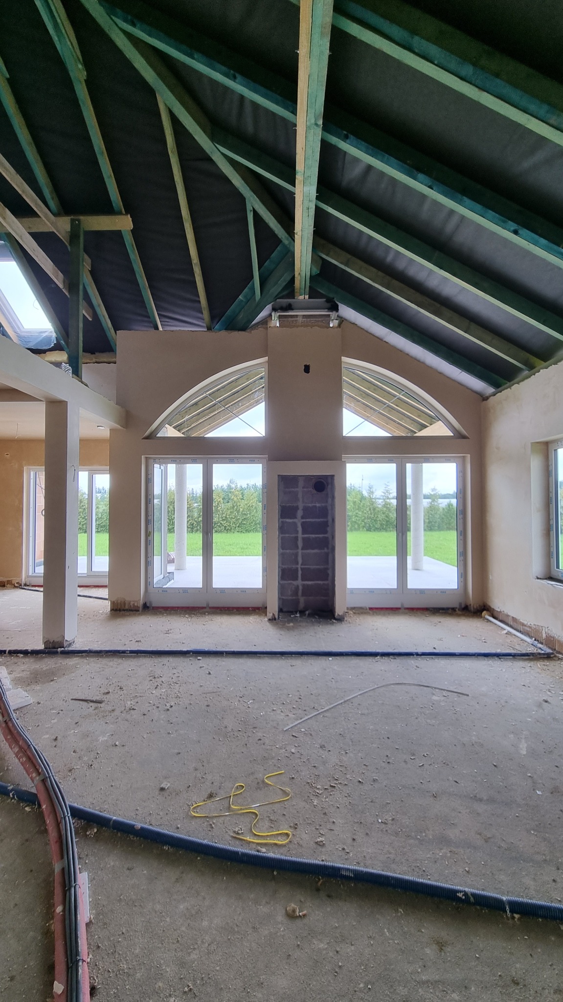 Wnętrze domu w trakcie prac wykończeniowych: ściany po nałożeniu pierwszej warstwy tynku, widoczne instalacje na podłodze, surowa konstrukcja dachu z izolacją.