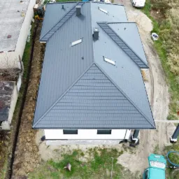 Widok z drona na dom z nowym, grafitowym dachem z dachówki ceramicznej, widoczne okna dachowe i komin, obok budynek gospodarczy i wykop pod fundamenty.