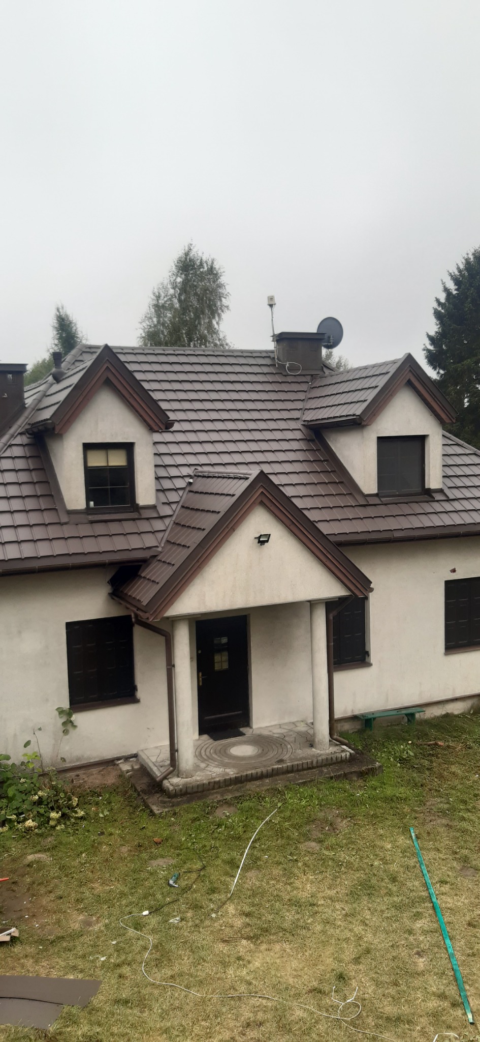 Dom jednorodzinny z nowym, brązowym dachem z dachówki ceramicznej, widoczne obróbki blacharskie, komin z anteną satelitarną, pochmurne niebo.