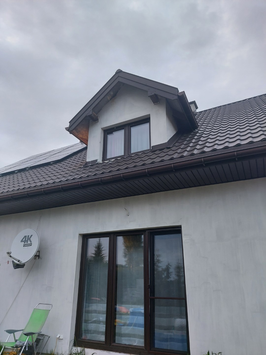 Dom jednorodzinny z brązowym dachem z dachówki trapezowej, nowymi oknami dachowymi i orynnowaniem, widok z dołu na szarym tle nieba.