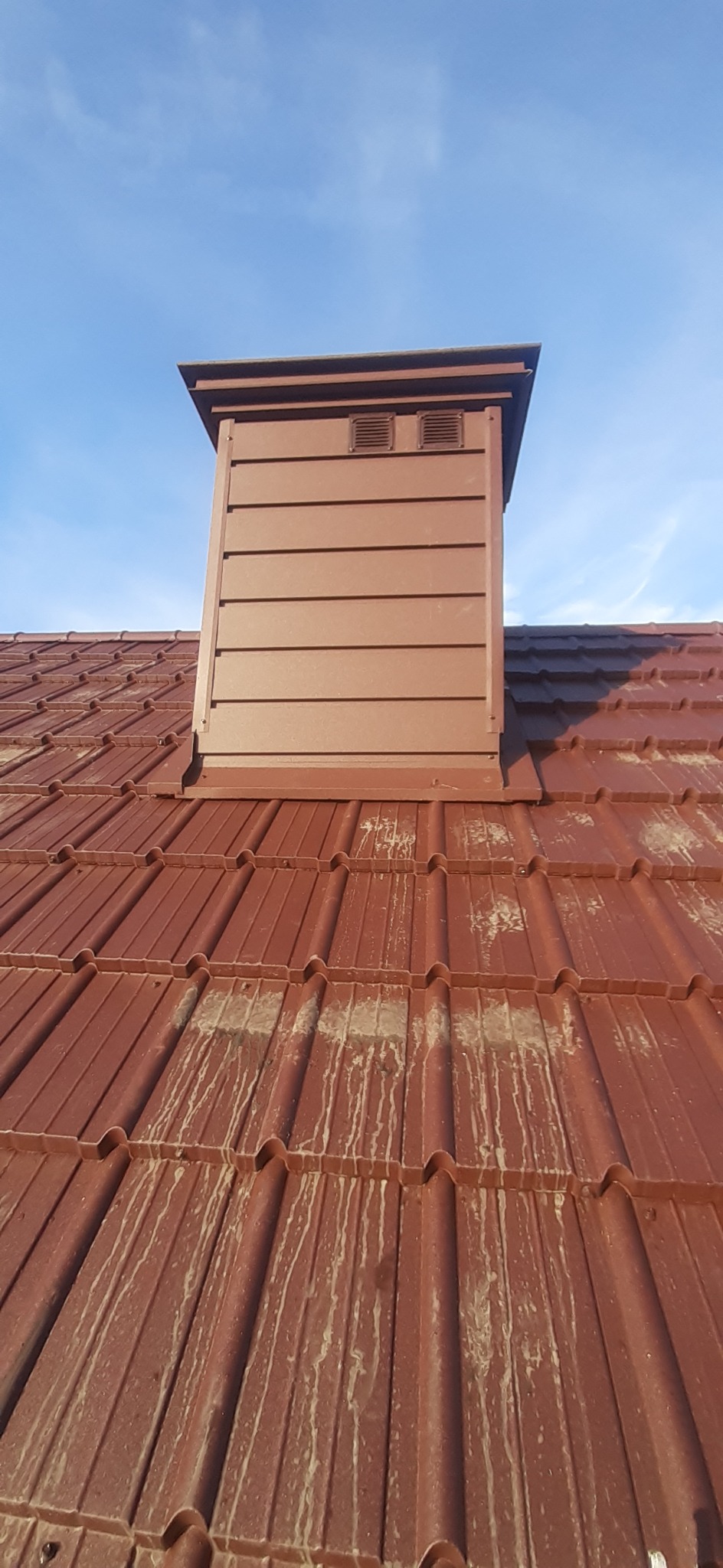 Czerwona dachówka ceramiczna z kominem w kolorze, widoczne zabrudzenia na powierzchni dachówek, błękitne niebo w tle.