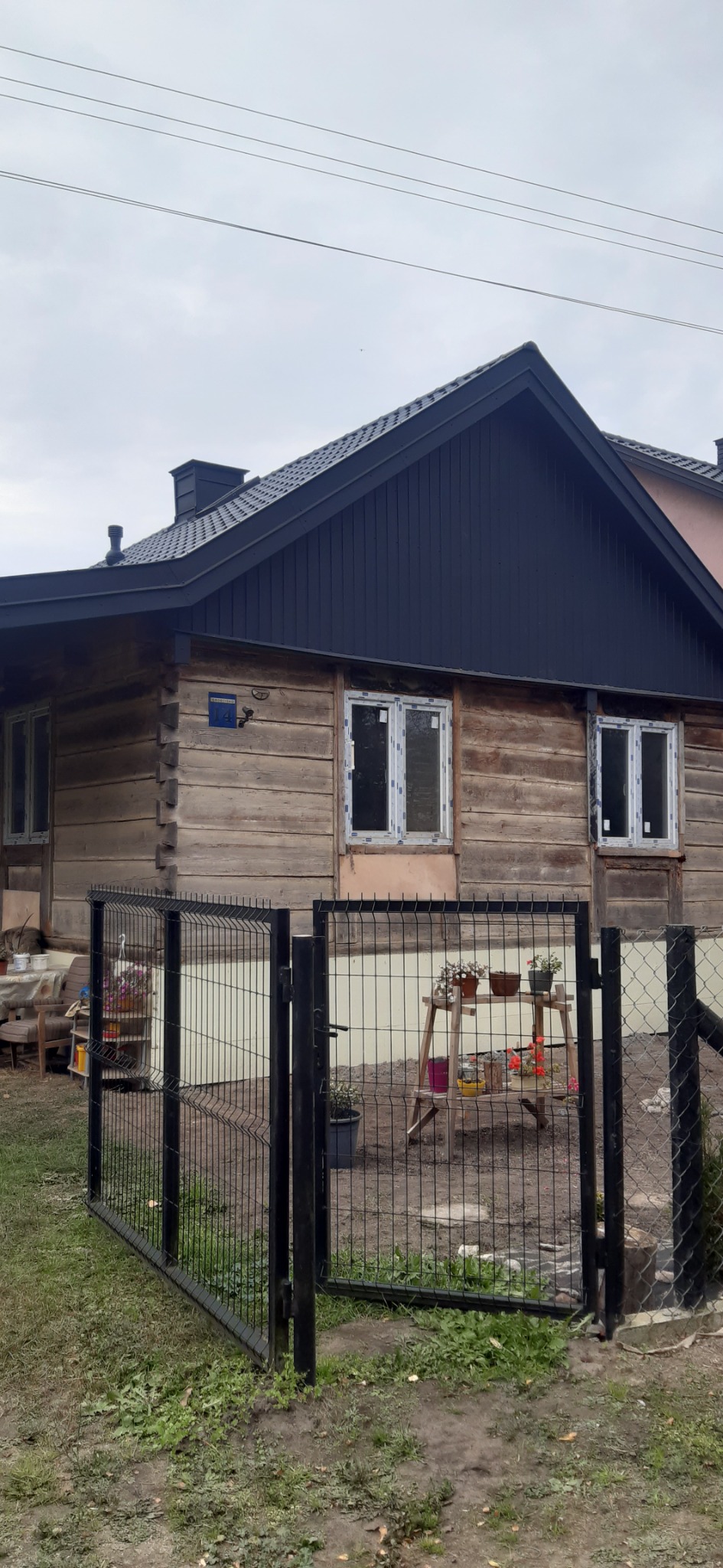 Dom z drewnianych bali z nowym, ciemnym dachem i nowymi oknami, ogrodzony czarnym metalowym płotem.