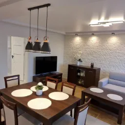 Salon z białymi ścianami o fakturze kamienia, ciemnymi meblami i nowoczesnym oświetleniem: wiszące lampy nad stołem i listwa LED na suficie.