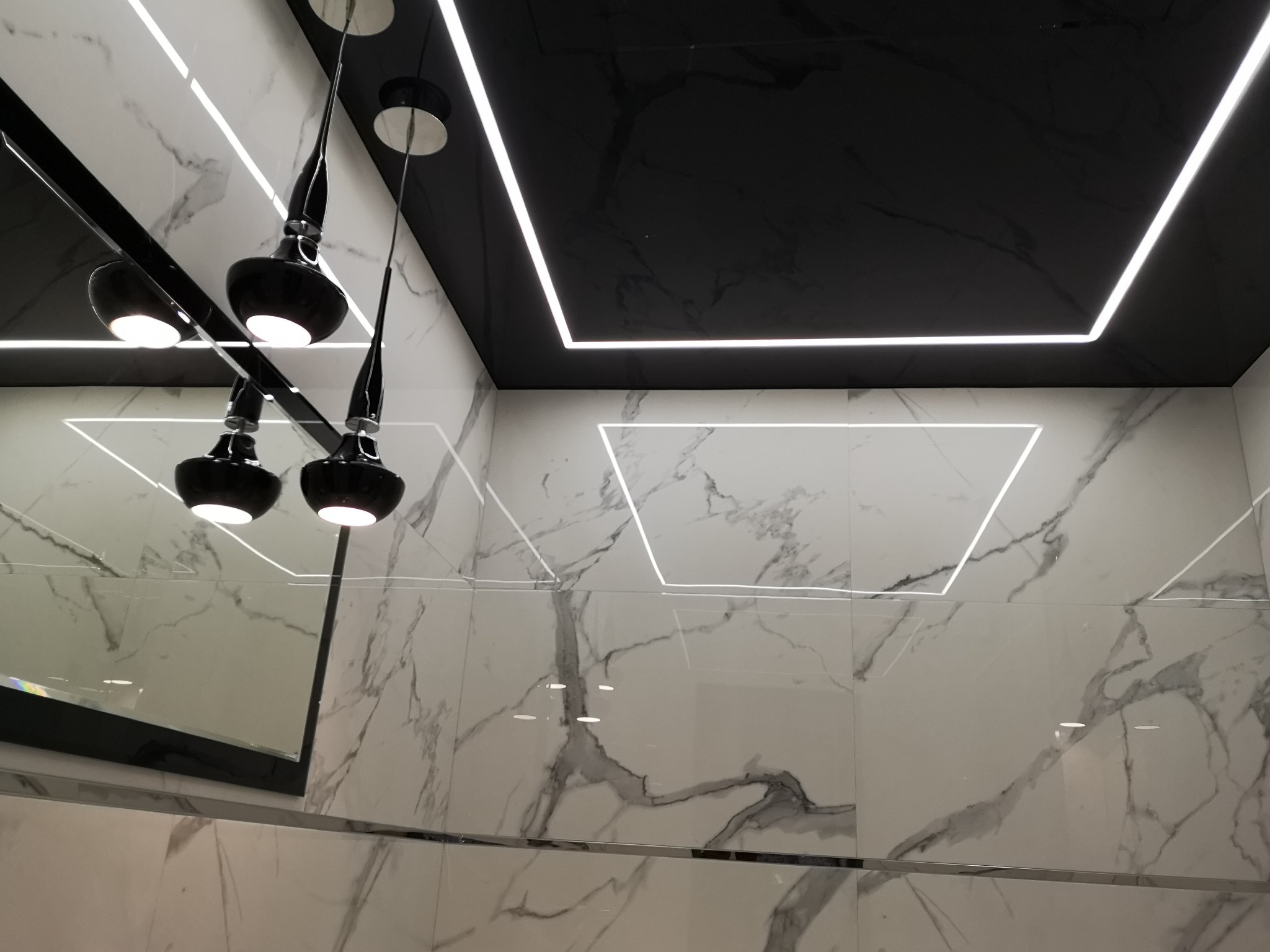 Nowoczesna łazienka z białymi płytkami imitującymi marmur, czarnym sufitem podwieszanym z oświetleniem LED i designerskimi czarnymi lampami wiszącymi, odbicie w lustrze.