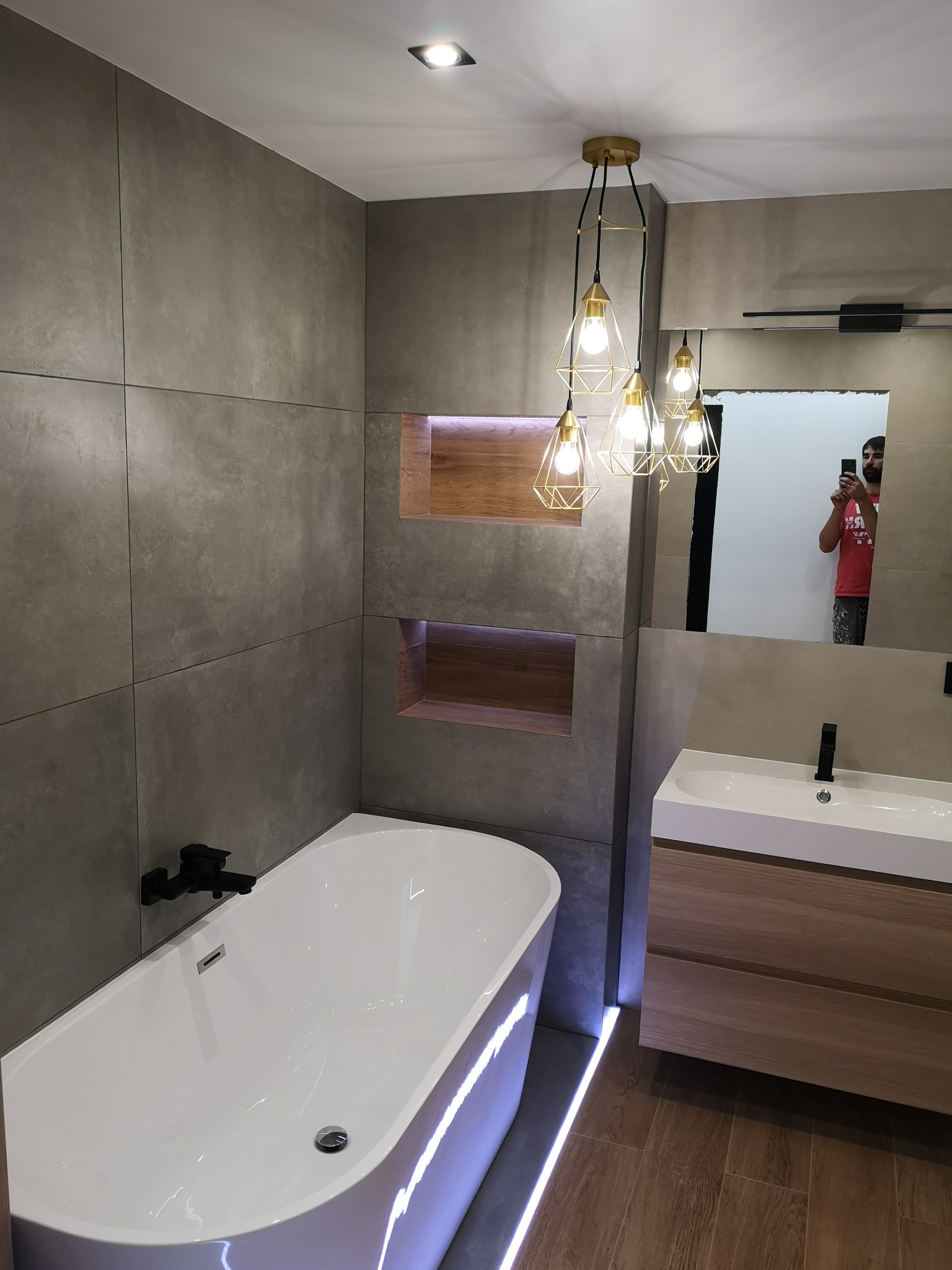 Nowoczesna łazienka z szarymi płytkami wielkoformatowymi, podświetlaną wanną, drewnianymi wnękami i złotą lampą wiszącą o geometrycznym designie, odbicie fotografa w lustrze.
