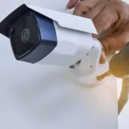 Instalacja kamery monitoringu: mężczyzna z wiertarką montuje białą kamerę na ścianie, widoczny zegarek na nadgarstku.