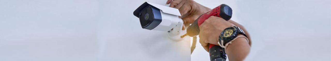 Instalacja kamery monitoringu: mężczyzna z wiertarką montuje białą kamerę na ścianie, widoczny zegarek na nadgarstku.