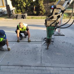 KSM HYDRO Żek Katarzyna - Dwóch pracowników w kamizelkach odblaskowych mierzy i wyznacza obszar do wykopu na betonowej nawierzchni ulicy za pomocą miarki i sprayu, obok stoi ciężki młot pneumatyczny na statywie.