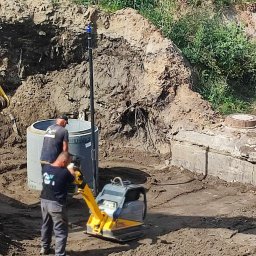 KSM HYDRO Żek Katarzyna - Prace ziemne: ubijanie gruntu zagęszczarką w wykopie obok betonowego zbiornika i studni, widoczna koparka i agregat pompowy.