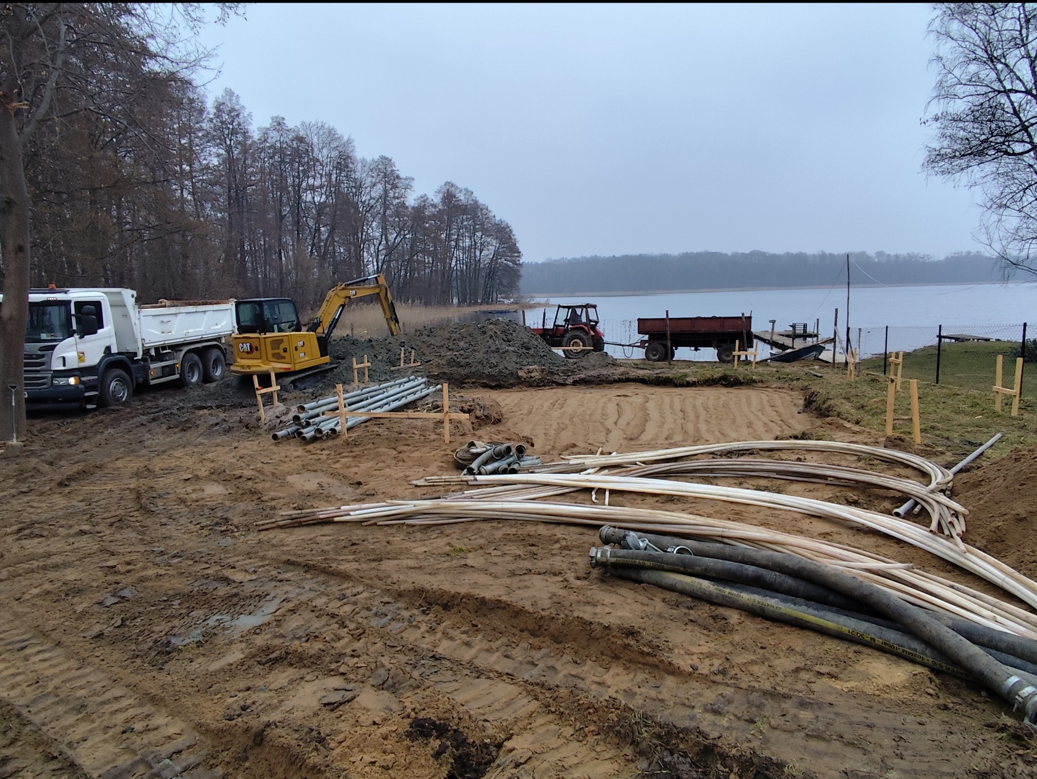 Teren nad jeziorem w trakcie przygotowania pod inwestycję; widoczne maszyny budowlane: wywrotka, koparka, traktor z przyczepą; rury różnego rodzaju przygotowane do instalacji.