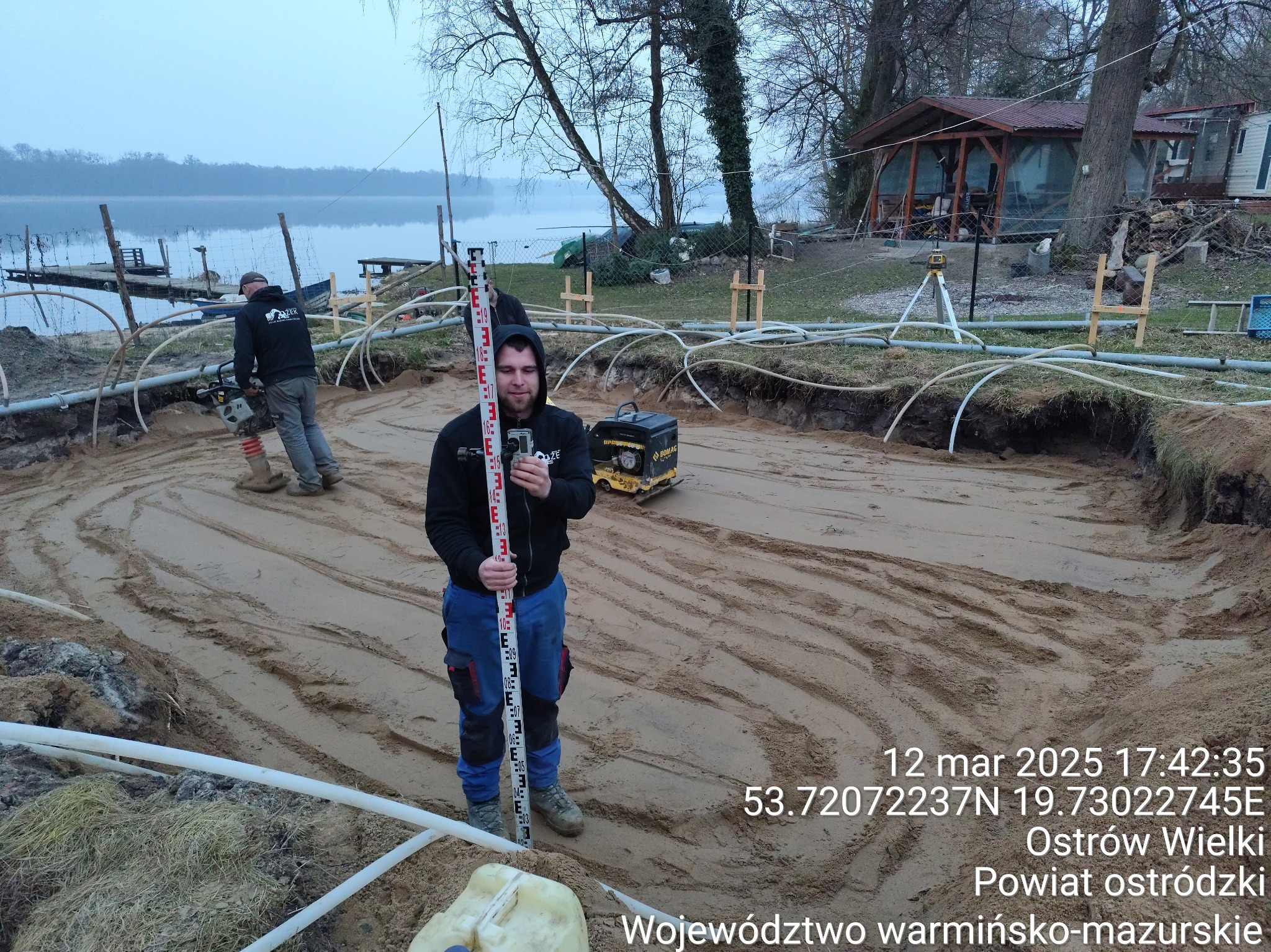 Pracownik z łatą niwelacyjną na tle wykopu z ułożonymi rurami, ubijarka gruntu i agregat prądotwórczy w tle, jezioro i zabudowania w oddali.