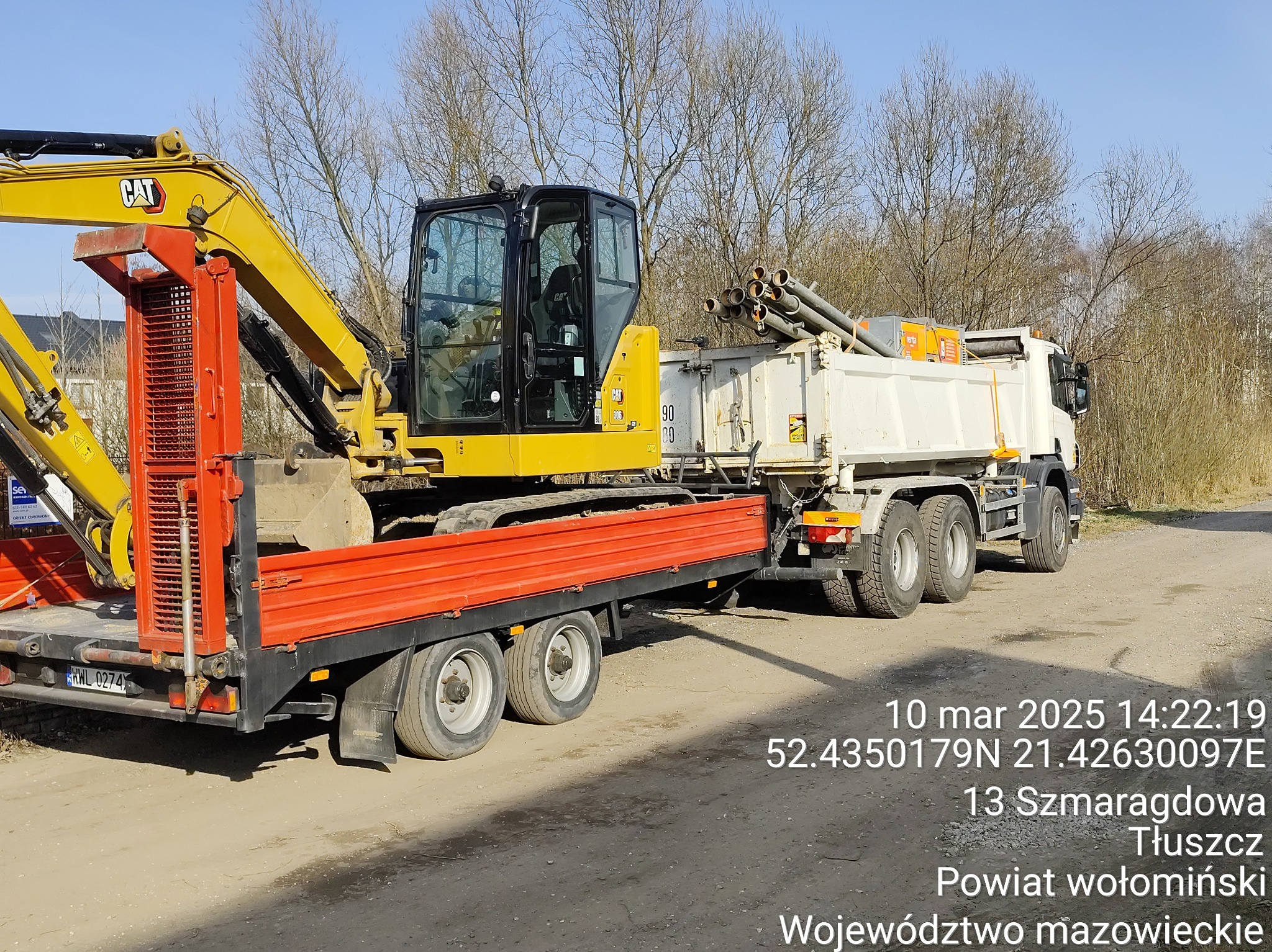 Żółta koparka CAT na czerwonej lawecie, za nią biała ciężarówka z rurami, w tle drzewa bez liści, na dole widoczny adres i data.