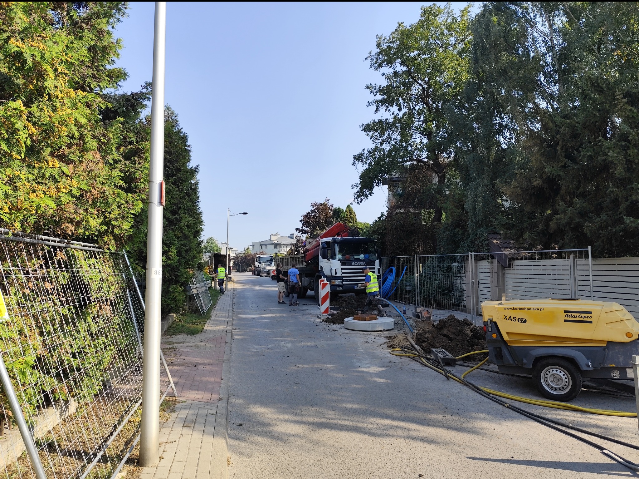 Ekipa budowlana przy wykopie na ulicy, ciężarówka Scania z dźwigiem, żółty kompresor Atlas Copco XAS 67, widoczne zabezpieczenia i narzędzia.