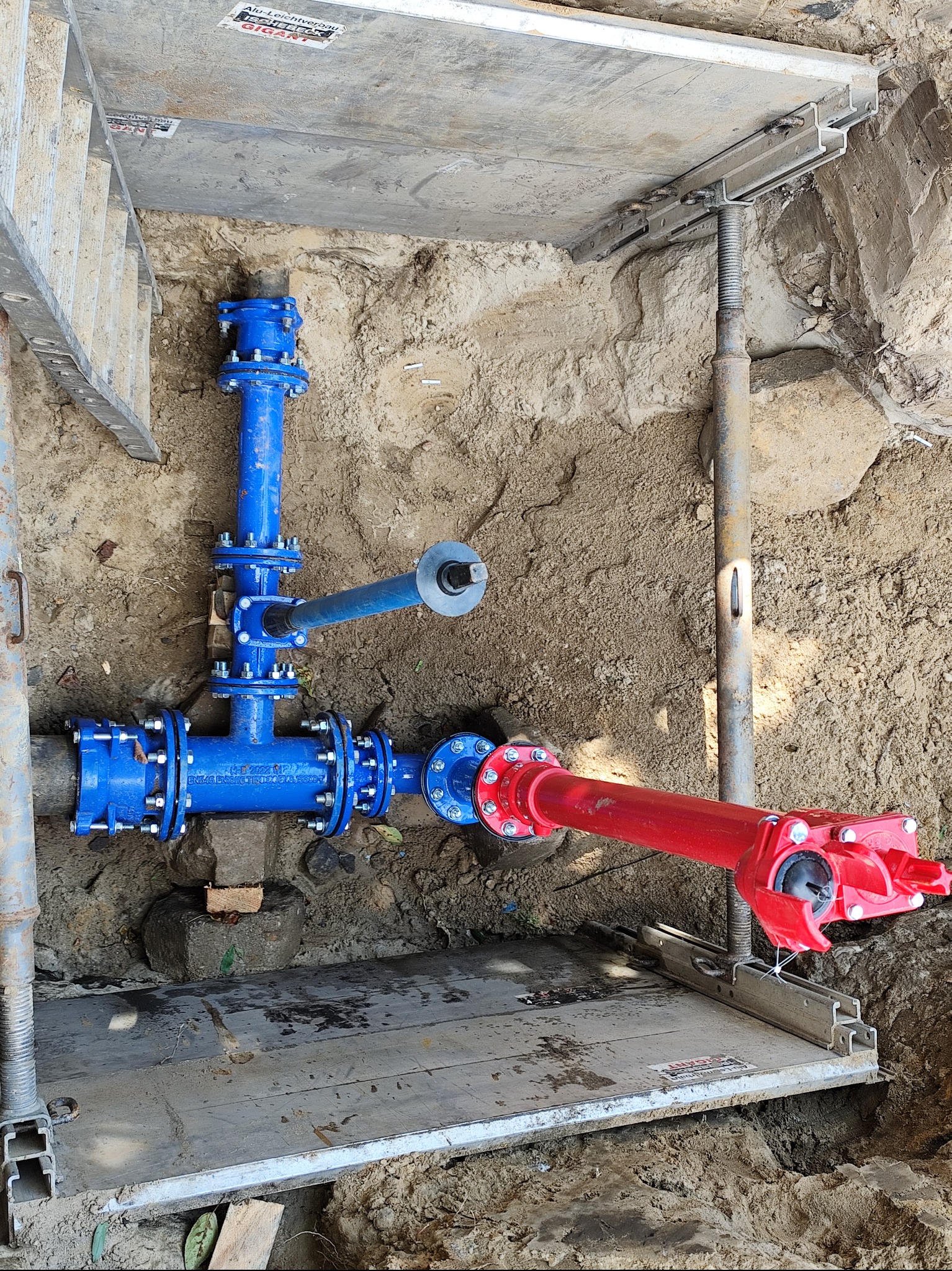 Wykonane w wykopie połączenie rur wodociągowych: niebieskie rury z zaworami łączą się z czerwoną rurą, zabezpieczone deskami i metalowymi stemplami.