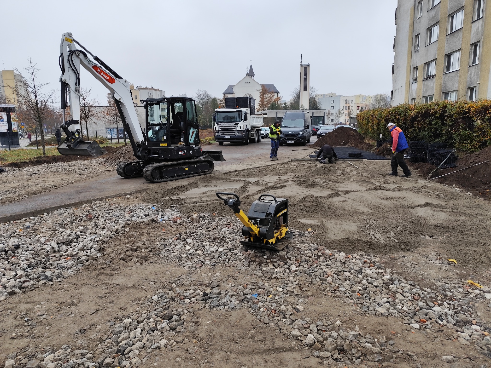 Prace ziemne z użyciem minikoparki Bobcat E60 i zagęszczarki gruntu, przygotowanie terenu pod budowę z widocznymi pracownikami w kamizelkach odblaskowych i transportem w tle, w oddali budynek...