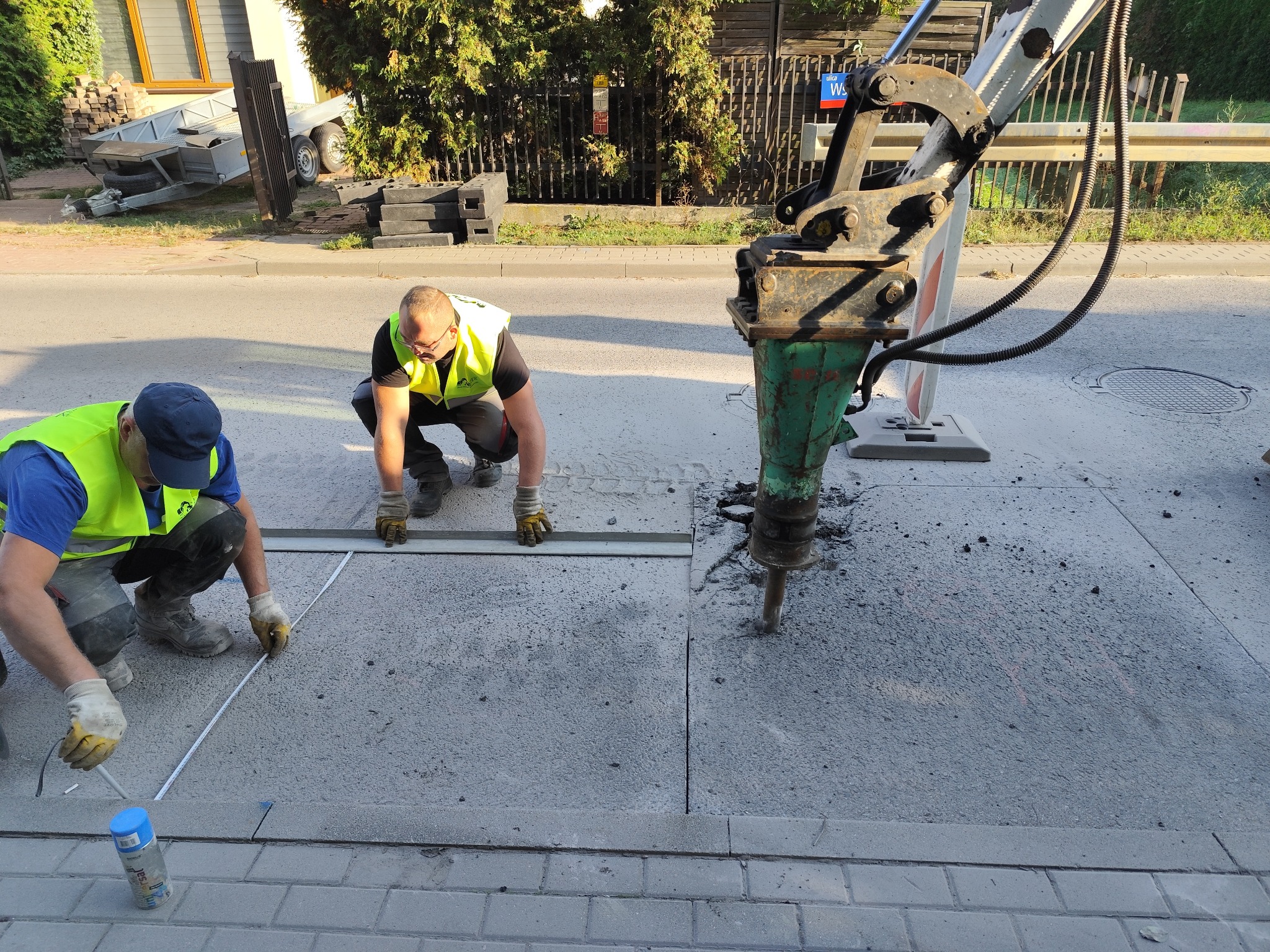 Dwóch pracowników w kamizelkach odblaskowych mierzy i wyznacza obszar do wykopu na betonowej nawierzchni ulicy za pomocą miarki i sprayu, obok stoi ciężki młot pneumatyczny na statywie.