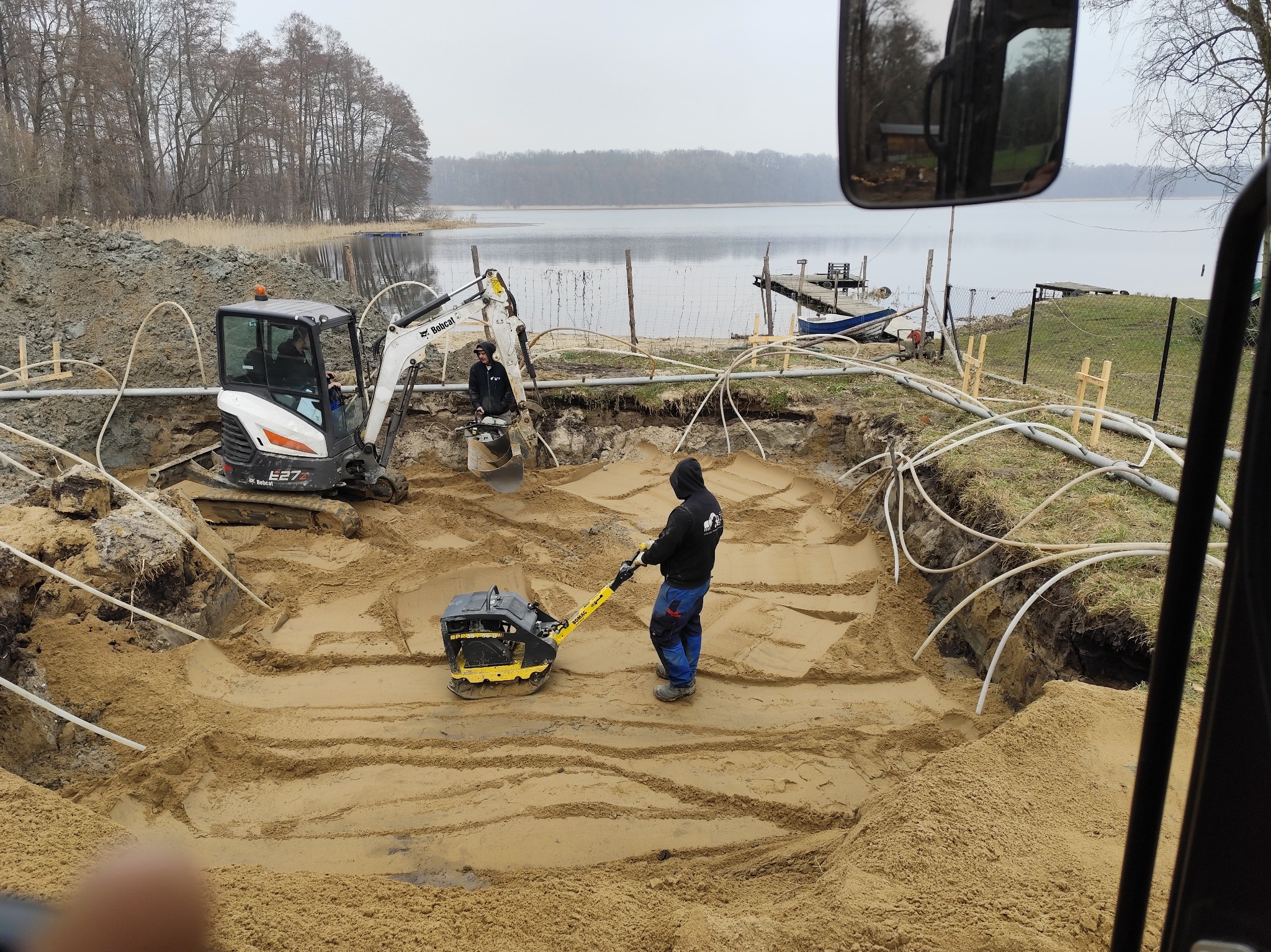 Prace ziemne przy budowie systemu rozsączania ścieków: operator zagęszczarki ubija piasek w wykopie, w tle minikoparka Bobcat i jezioro z pomostem.