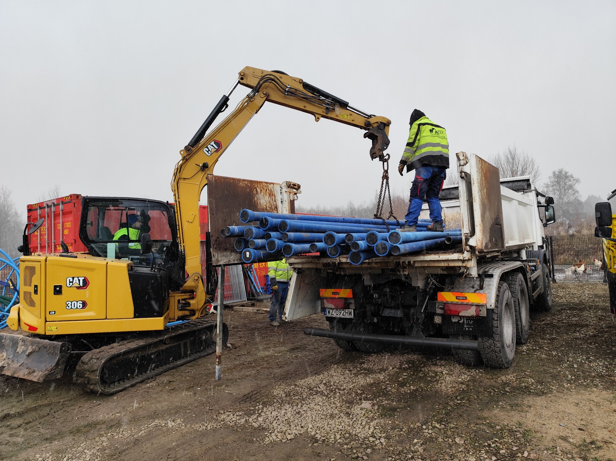 Żółta koparka CAT 306 obok ciężarówki z niebieskimi rurami, pracownik w odblaskowej kurtce na pace ciężarówki, drugi pracownik obok koparki, teren budowy w pochmurny dzień.