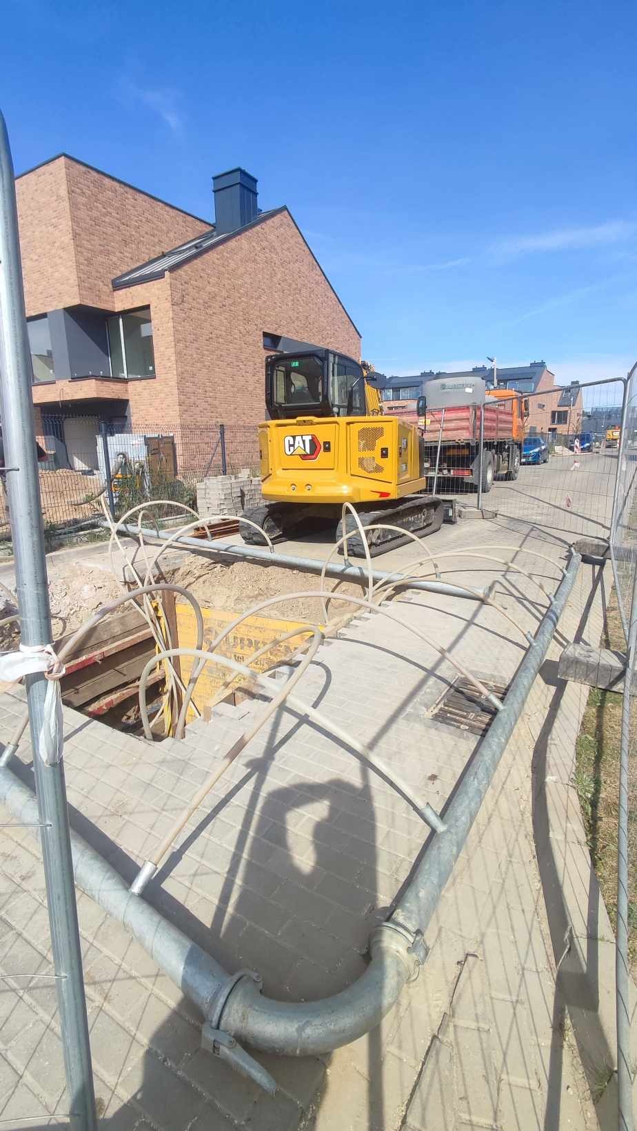 Żółta koparka CAT na gąsienicach stoi przy wykopie zabezpieczonym metalowymi szalunkami, w tle budynek mieszkalny i wywrotka.