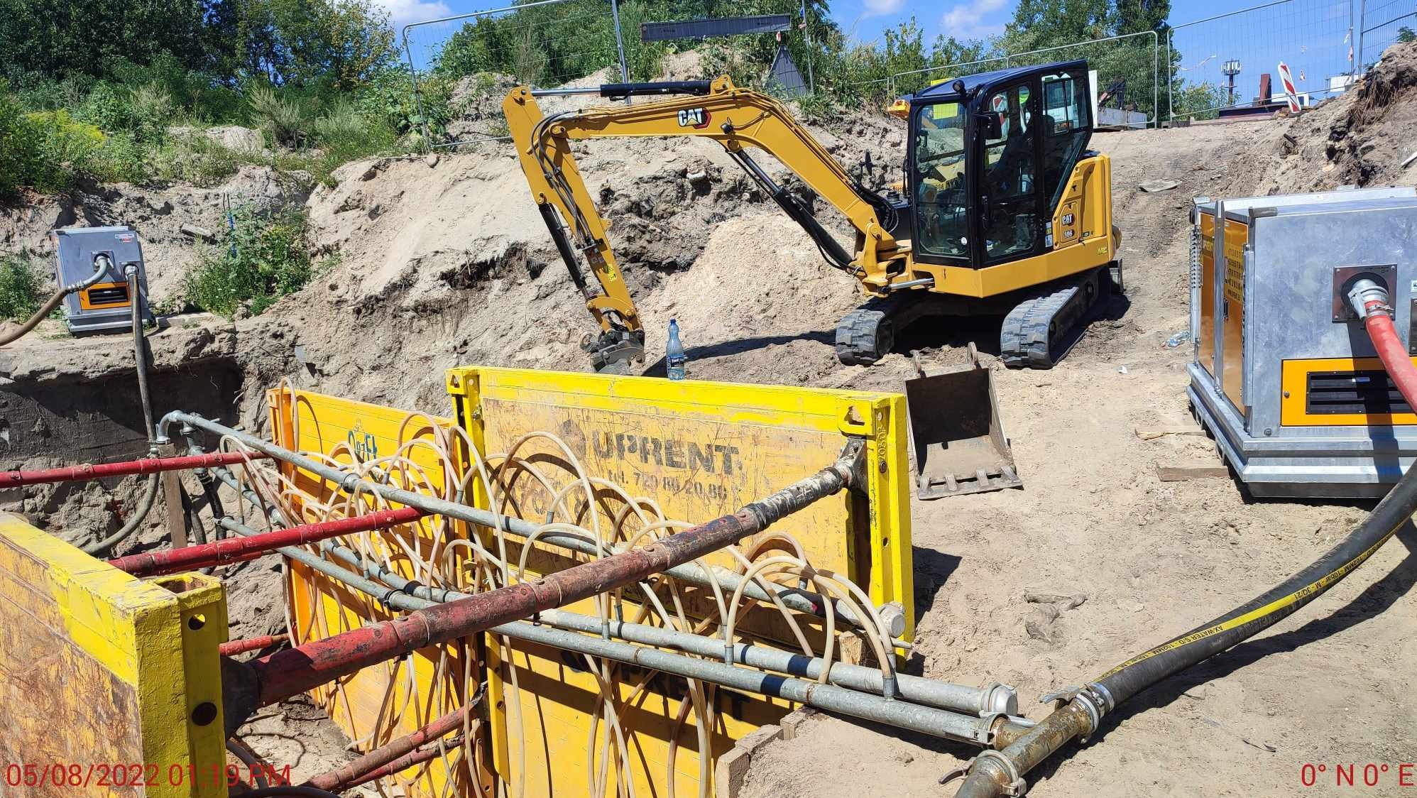 Żółte szalunki wykopu z rurami i pompami odwadniającymi, koparka CAT w tle, teren budowy.