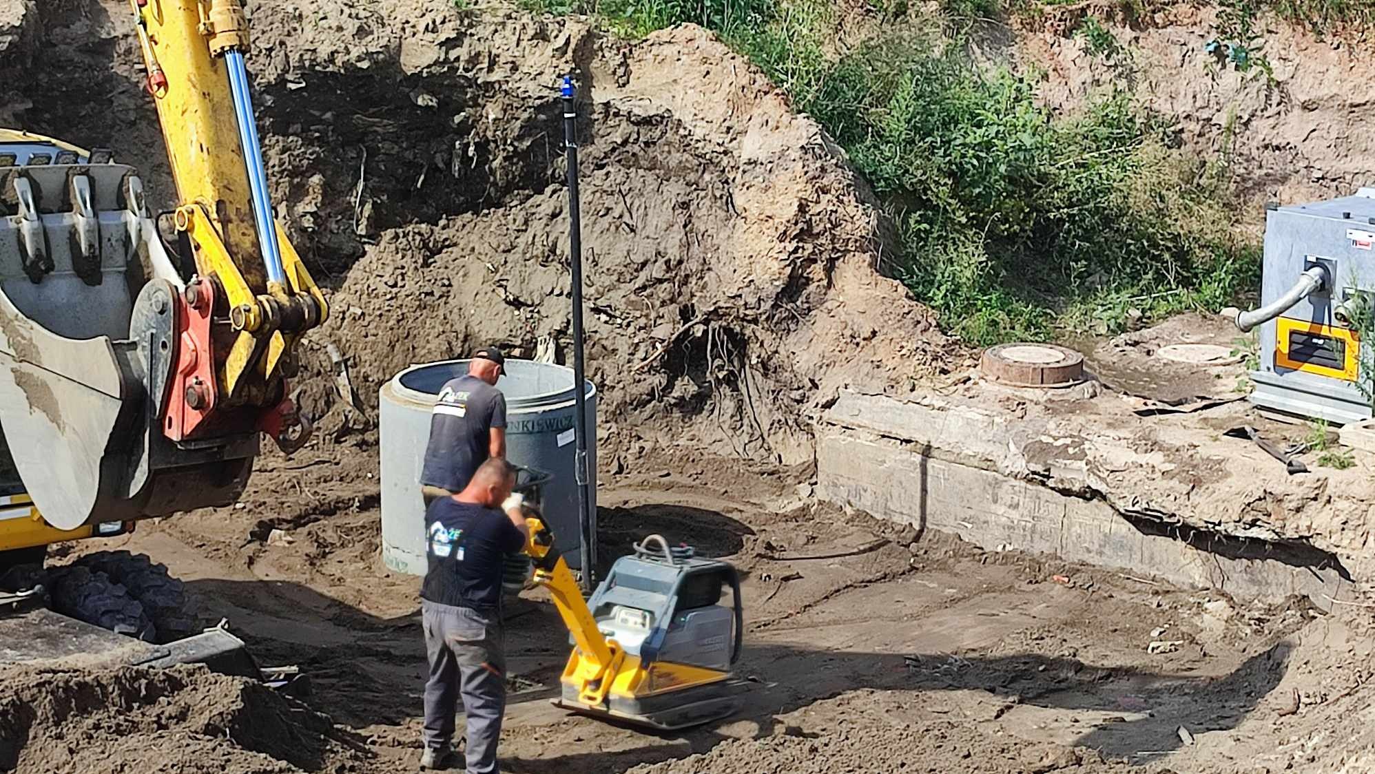 Prace ziemne: ubijanie gruntu zagęszczarką w wykopie obok betonowego zbiornika i studni, widoczna koparka i agregat pompowy.