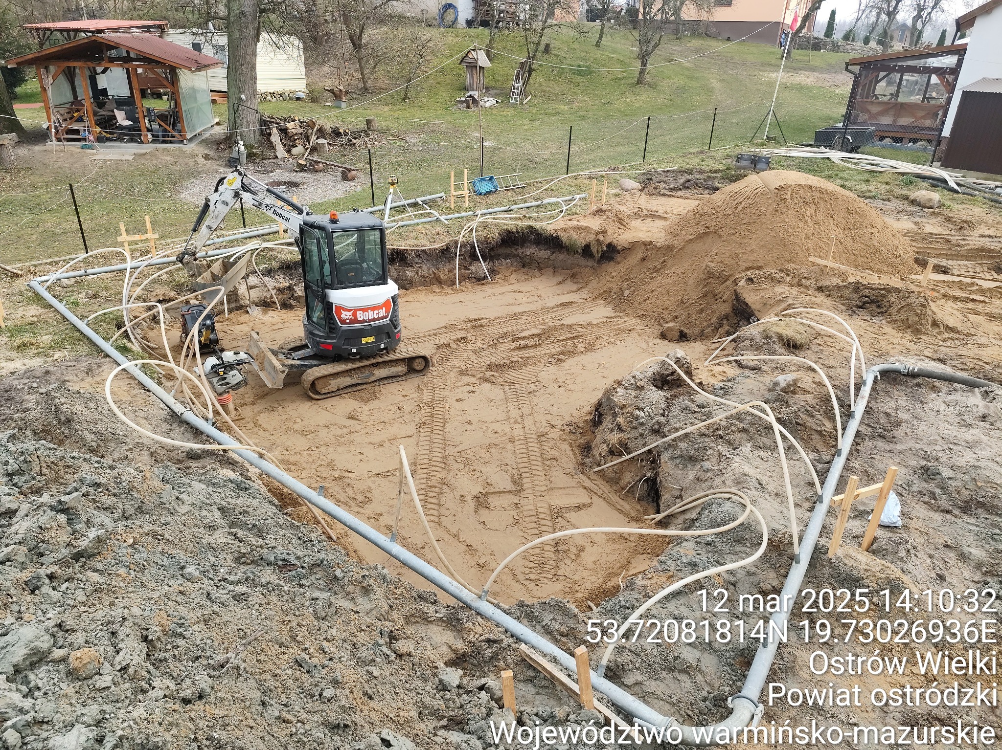Wykonanie wykopu pod instalację sanitarną z użyciem minikoparki Bobcat. Widoczne rury i białe przewody na dnie wykopu. Koordynaty GPS i data na zdjęciu.
