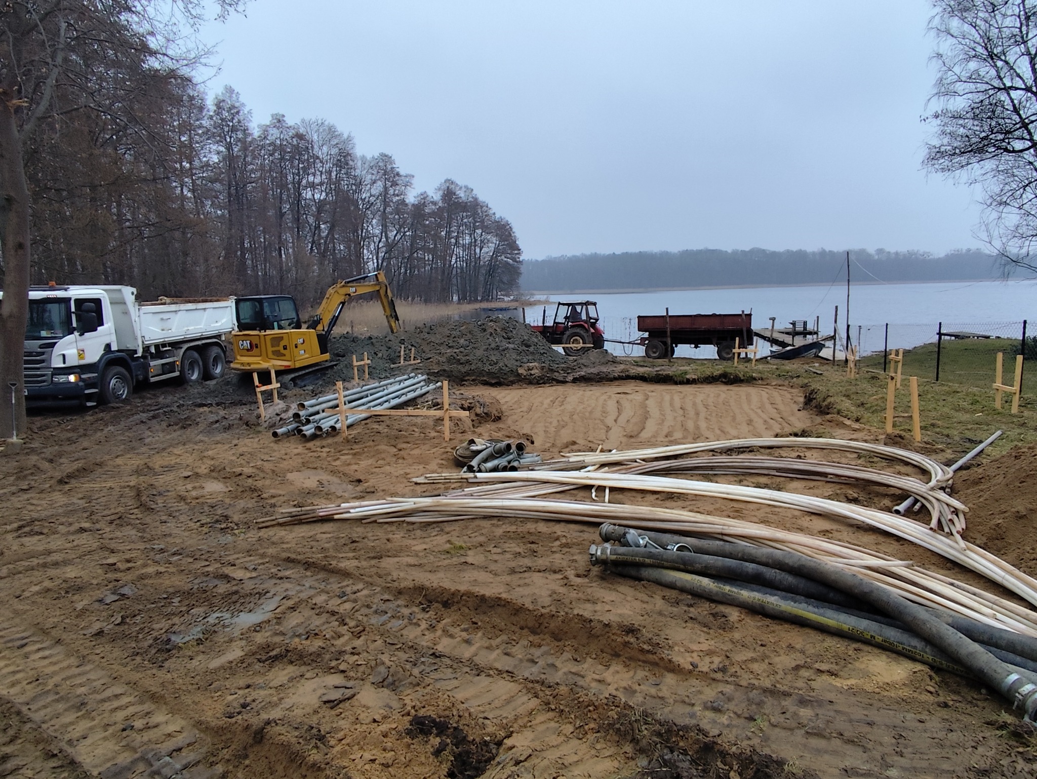 Prace ziemne nad jeziorem: wykop, rury, ciężarówka i koparka CAT. W tle traktor z przyczepą i pomost. Pochmurne niebo.
