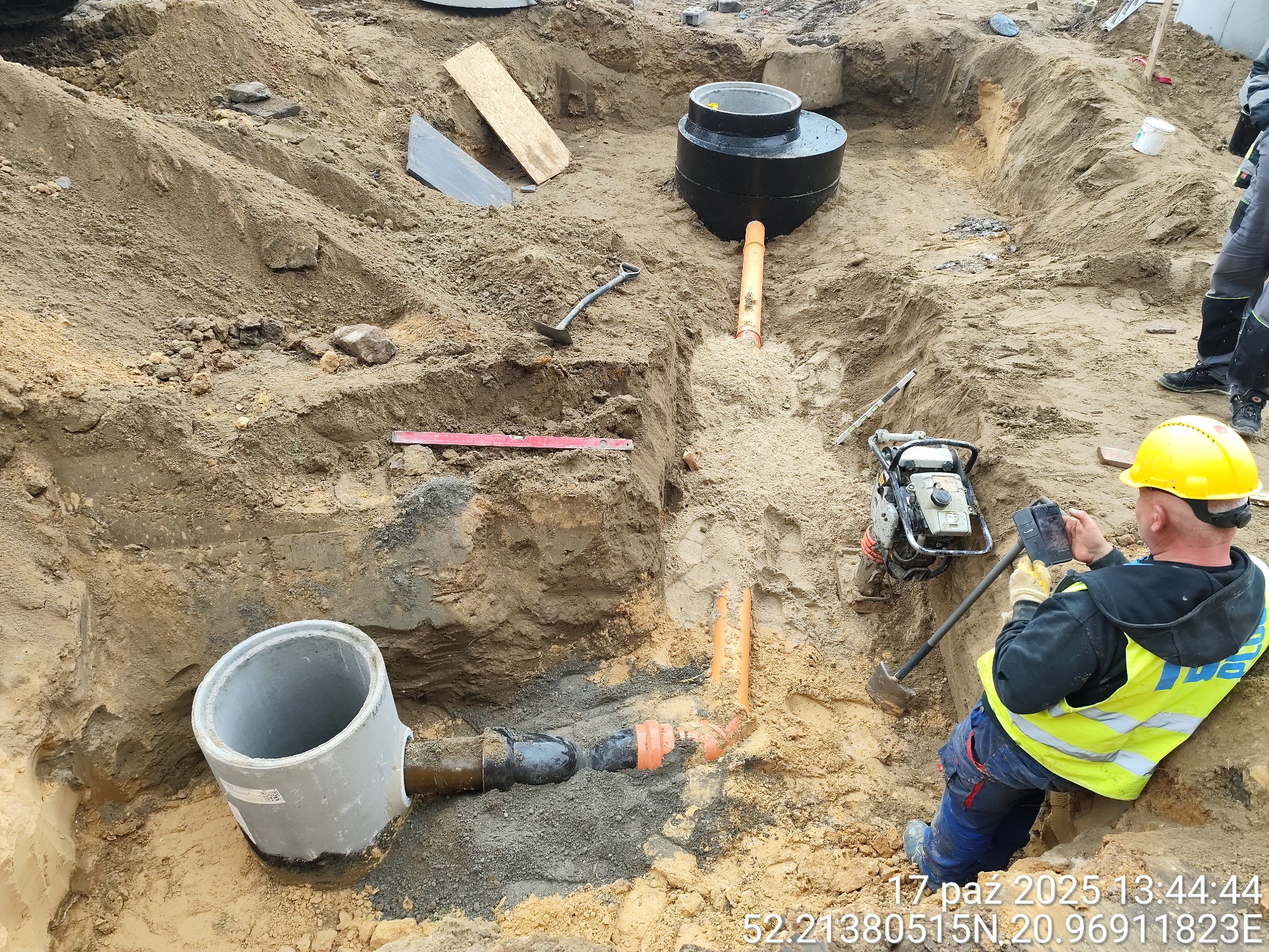 Montaż szamba betonowego w wykopie. Widoczne elementy instalacji, rury, studzienka betonowa i pracownik w kasku dokumentujący postęp prac.