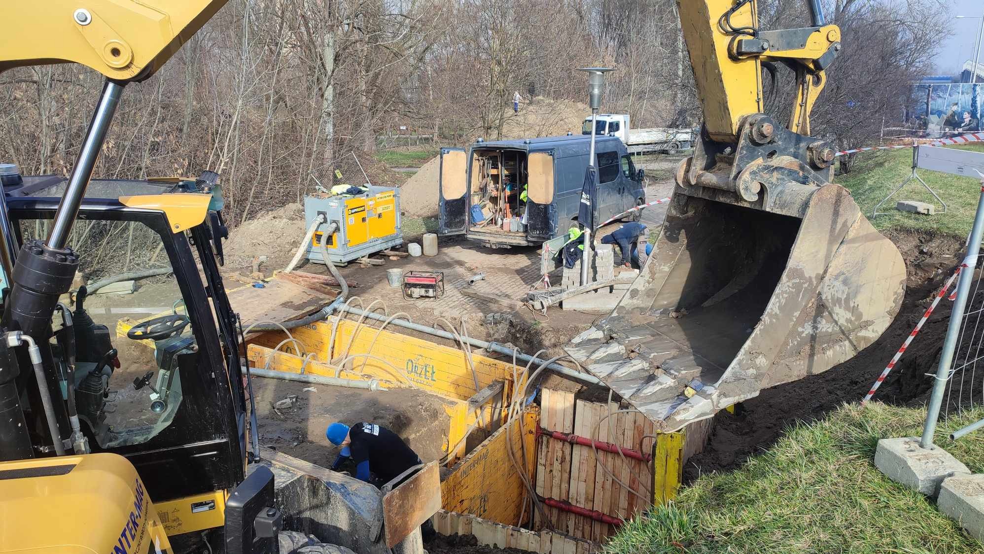 Żółty wykop zabezpieczony szalunkiem stalowym, z pracującymi robotnikami i sprzętem budowlanym w tle, sugerujący etap prac przyłączeniowych.