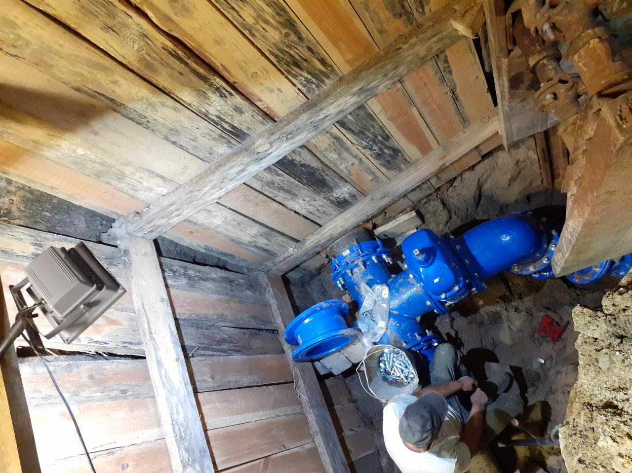 Montaż niebieskich rur wodociągowych w wykopie z drewnianym szalunkiem, widoczny pracownik z wiadrem pełnym śrub i lampa oświetlająca miejsce pracy.